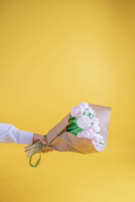 A person wearing a white shirt holds a bouquet of soft pink tulips wrapped in light brown kraft paper with green ribbon, presented against a bright yellow background. The bouquet features fresh tulips