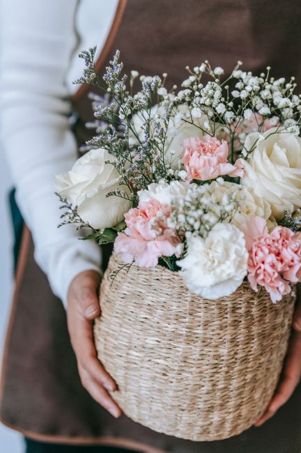 A person holding a round woven wicker basket filled with a floral arrangement featuring white roses, soft pink carnations, delicate sprigs of baby's breath, and lavender accents, all arranged in a lus