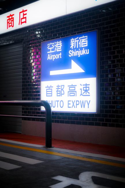 The image features a large illuminated directional sign at an airport, with the sign indicating directions to Shinjuku and the Shuto Expressway in both Japanese and English. The sign is mounted on a b