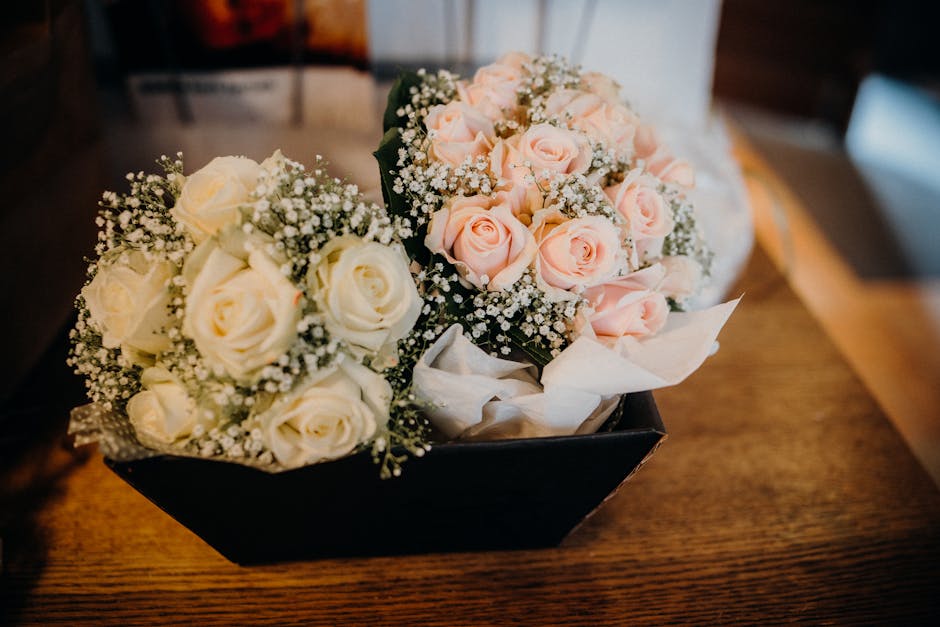 A gift of two floral bouquets placed in a black rectangular box sits atop a wooden surface. One bouquet features cream-colored roses with delicate petals and tiny clusters of baby's breath, wrapped pa