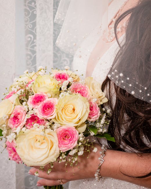 A bride holds a round bridal bouquet featuring white roses, pale pink lisianthus, and soft purple and green accents, wrapped with a pink ribbon. The bouquet displays fresh, velvety petals and green fo