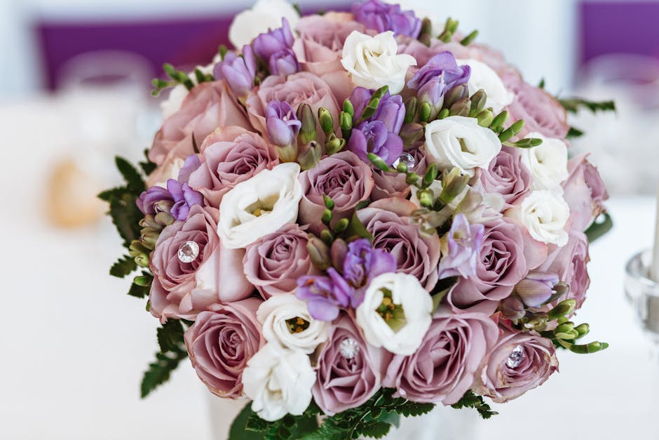 A round floral bouquet featuring soft pink roses, white lisianthus, and light purple freesias, arranged with green foliage and accented with small rhinestone embellishments. The flowers are fresh, wit