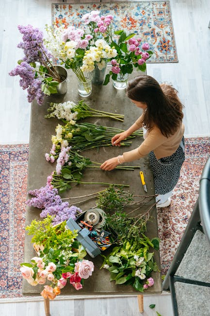 A woman with long brown hair, wearing a beige top and patterned trousers, arranges fresh floral stems behind a large wooden table in a well-lit indoor space. The table is covered with various types of