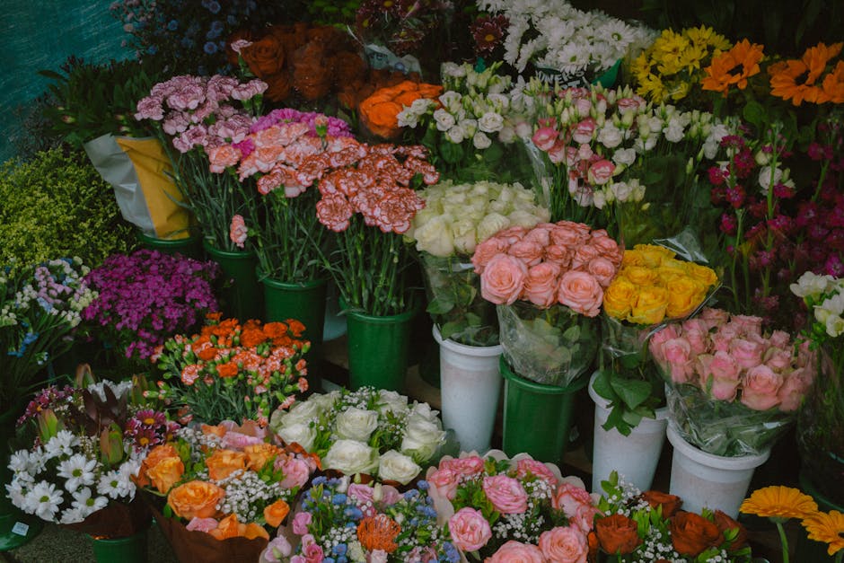 A vibrant display of fresh floral arrangements and bouquets arranged in green and white pots and wrapping, featuring a variety of flowers including pink and peach roses, white chrysanthemums, purple a